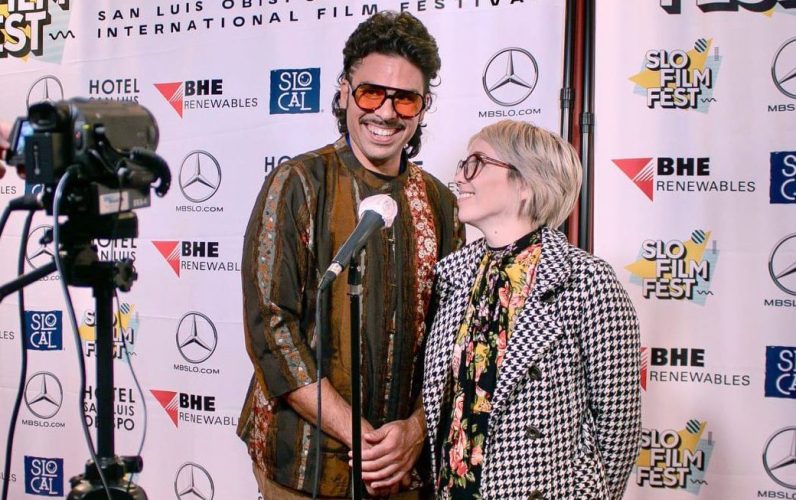 One male filmmaker and one female filmmaker standing in front of a mic and camera on the red carpet and smiling.