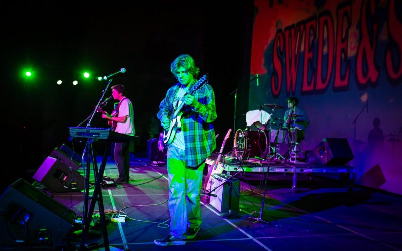 In the center of the frame a man plays an electric guitar with a keyboardist in the back and drummer on the right side of the stage.