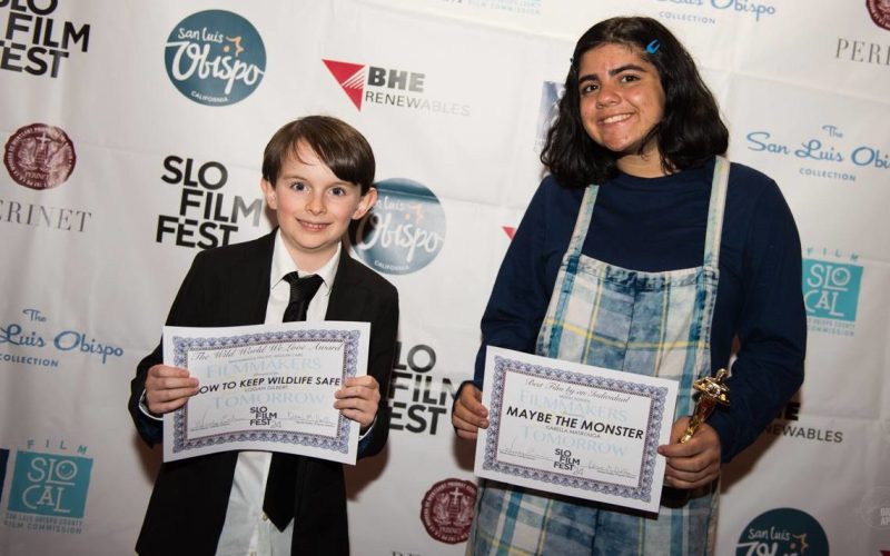 A young male filmmaker holds his award in his hand next to another female filmmaker as they both smile.